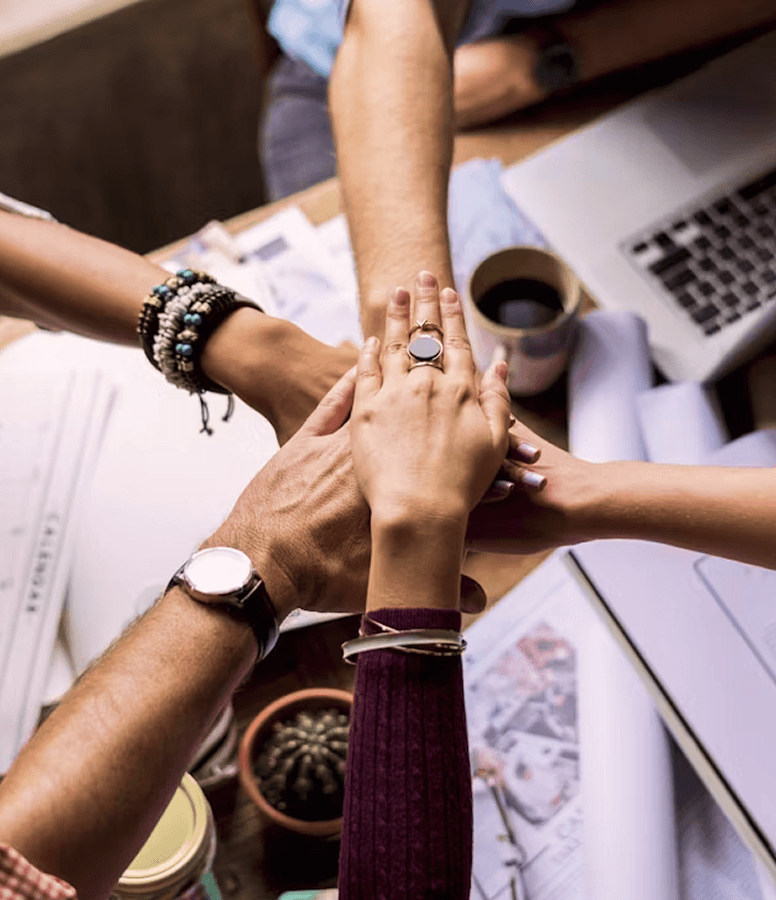 Hands stacked together in a teamwork huddle over a workspace.